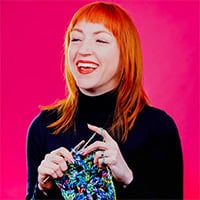 The top half of a woman with bright orange hair on a pink background, working on a multicolored project.