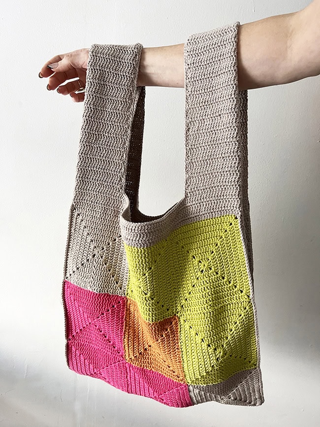 An arm held out with a crocheted bag hanging on it. The bag is mostly taupe with some blocks of color on the body.