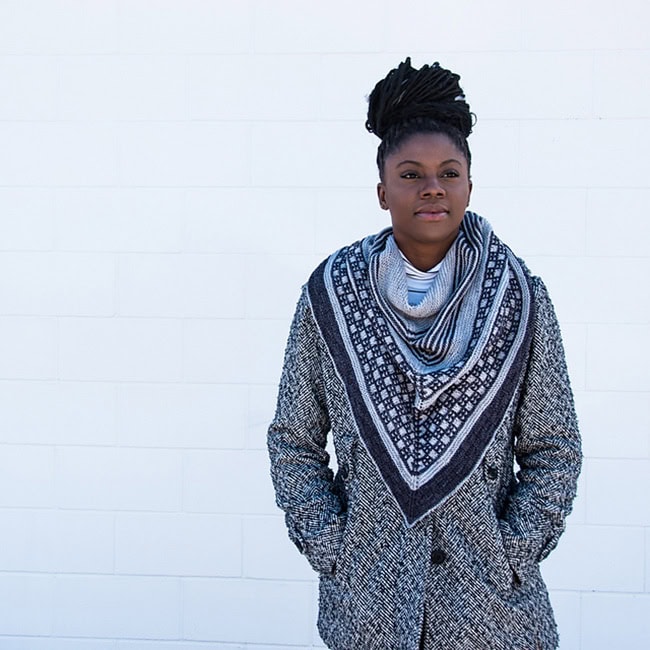 A Black woman wearing a knitted triangular shawl in blues. The shawl has stripes in varying shades and a section that is a grid of boxes.