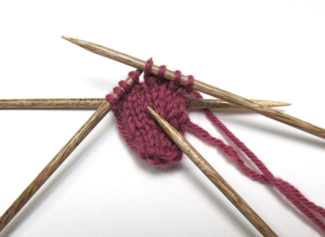 A set of 4 wooden double-pointed needles. There is a small-circumference rose-colored tube in the process of being knitted.